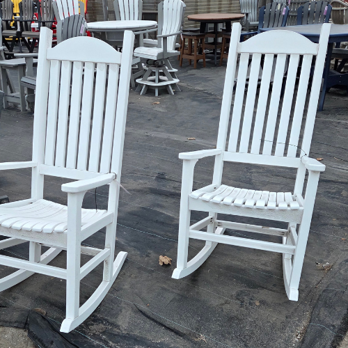 Two white wooden rocking chairs on a dark floor outdoors, surrounded by various patio furniture. The scene is calm and inviting, suggesting relaxation.