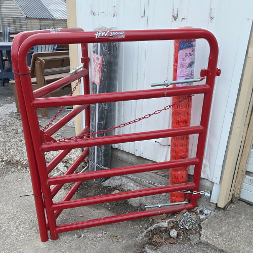 Red metal gate with a latch and chain, leaning against a white building. Nearby are orange mesh fencing rolls and wooden furniture. Industrial setting.