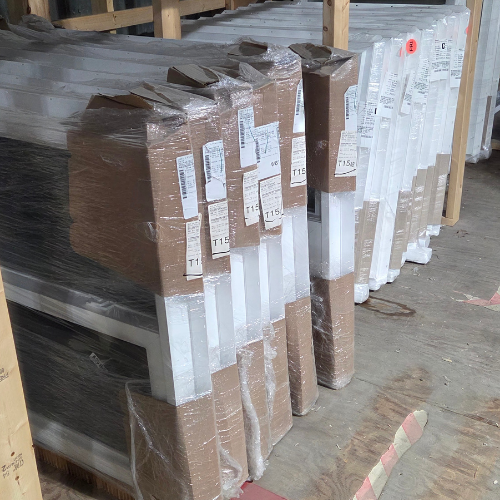 Rows of white windows are stacked vertically, wrapped in plastic with cardboard edges, and set on a wooden floor in a storage area. Labels are visible.