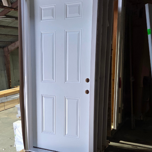 A white six-panel door stands upright in a storage area, surrounded by wooden beams and materials. The setting is dimly lit, giving a workshop ambiance.