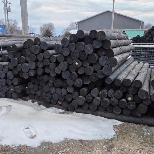 A large stack of black, cylindrical utility poles is piled neatly on a snowy and muddy ground. In the background, a utility building is visible under a cloudy sky.
