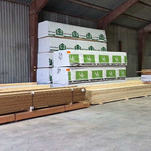 Stacks of lumber and plywood are neatly arranged inside a large warehouse. The wooden beams and planks are wrapped in branded packaging, suggesting a setting ready for construction or supply distribution. The atmosphere is industrial and organized.