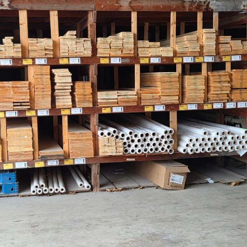 Stacks of wooden planks and PVC pipes are neatly organized on wooden shelves in a hardware store, suggesting a setting of order and readiness for building projects.