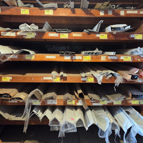 Wooden shelves stacked with various metal profiles wrapped in clear plastic, each compartment labeled with yellow tags. The scene is orderly and industrial.