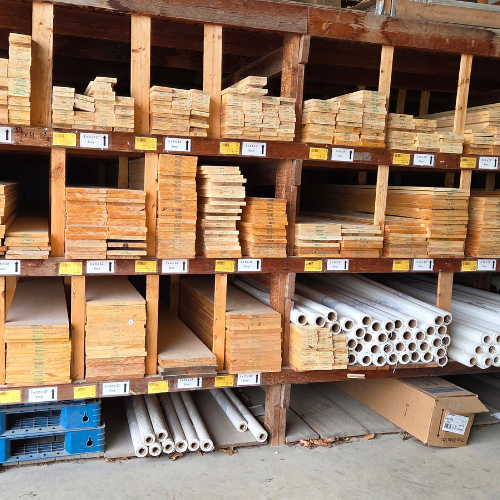 Wooden shelves filled with stacks of lumber and PVC pipes, organized and labeled in a hardware store aisle. The scene feels orderly and utilitarian.