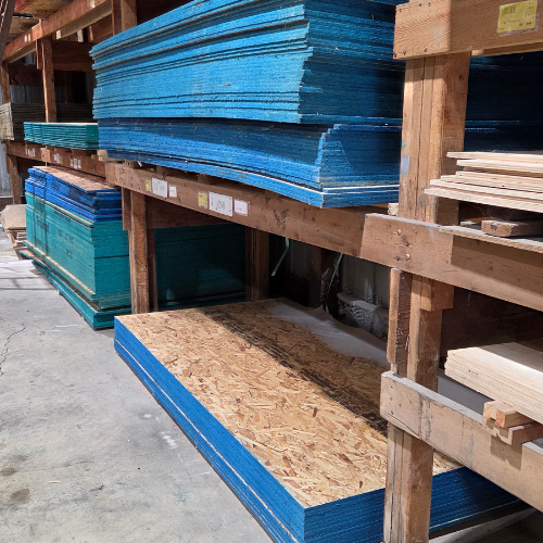 Shelves in a warehouse hold stacks of blue and turquoise foam boards and wooden panels. The scene is orderly and industrial with a calm tone.