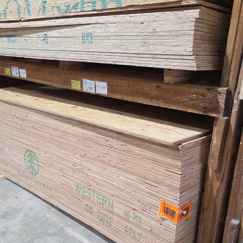 Stacks of plywood sheets are stored on wooden racks in a warehouse. The plywood has visible labels and barcodes, indicating organized inventory management.