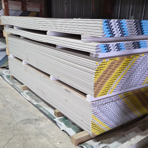 Stacks of drywall sheets arranged neatly on wooden pallets in a warehouse. The setting is industrial, with muted colors and a focus on organization.