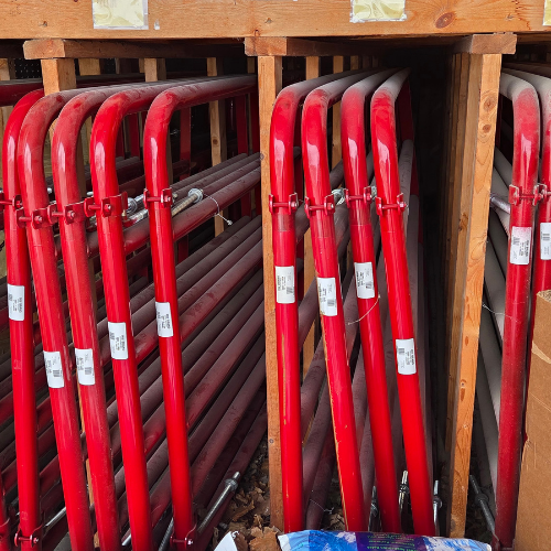 Red metal gate panels stacked vertically in a wooden storage area. The bright red color contrasts with the brown wood, creating a vibrant scene.