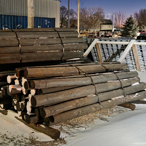 Stacked wooden logs tied in bundles are set on snowy ground outside a building. Metal racks are nearby, with a tree and sunset in the background.