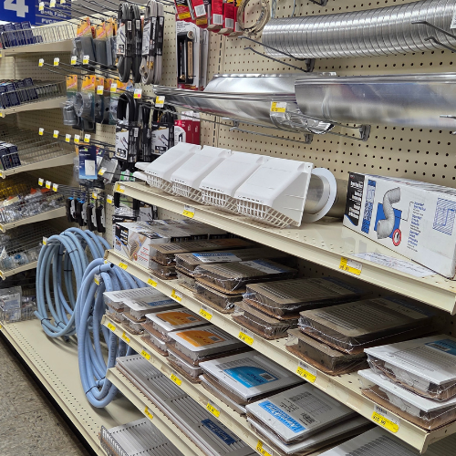 Hardware store aisle with shelves displaying air vent covers, aluminum ducts, hose pipes, and various tools. A practical, organized setting.