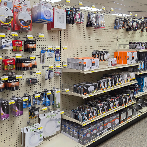 Store aisle with shelves stocked with various home electronics, including smoke detectors, batteries, and light fixtures, against a beige pegboard backdrop.