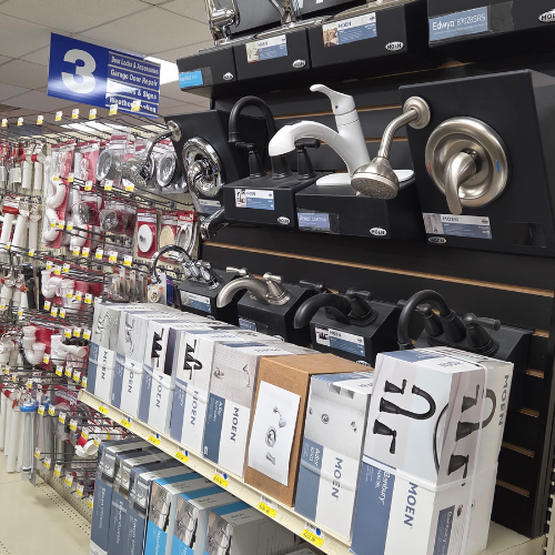 A hardware store aisle displays various metal faucets and showerheads on shelves. Boxes and packaged plumbing fixtures are neatly arranged below.
