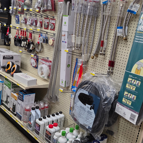 A hardware store aisle with shelves full of plumbing supplies, including stainless steel hoses, connectors, and bottles of cleaning products. Multicolored packaging adds vibrancy.