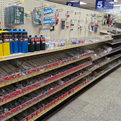 A hardware store aisle with shelves displaying bottles of propane, glue, light bulbs, and batteries. The aisle is organized and brightly lit.