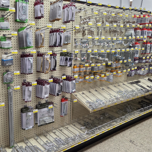 A store aisle with neatly arranged metal hardware items on a pegboard. Display includes handles, latches, and other small tools, all with price tags.