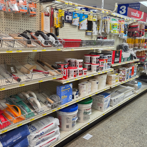A hardware store aisle displays paint supplies: brushes, rollers, and various paint buckets. The shelves are organized neatly with a wide color range.