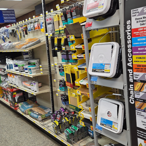 Hardware store aisle displaying various tools and supplies. Shelves hold paint cans, chains, and accessories. The atmosphere is organized and practical.