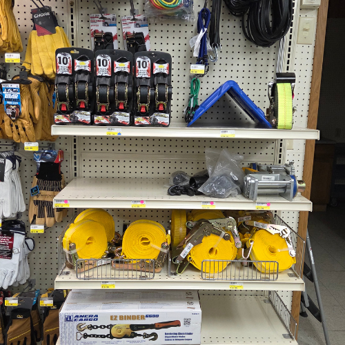 A store shelf displays various hardware supplies: yellow straps, ratchet tie-downs, gloves, and cables, conveying a utilitarian, organized tone.
