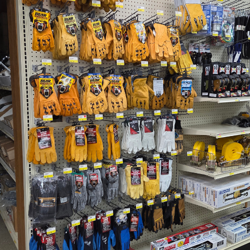 A hardware store display of assorted work gloves in various colors including orange, white, gray, and blue, alongside tools and tape.