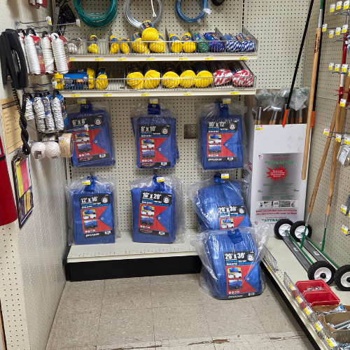Hardware store aisle displaying various blue tarps of different sizes on a shelf. Nearby shelves hold yellow balls, ropes, and gardening tools. Utilitarian feel.
