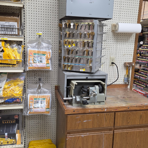 A hardware store corner with a key-cutting machine on a wooden counter. Above it hang rows of blank keys on a metal display. Nearby shelves hold packaged bags. The scene feels utilitarian and organized.
