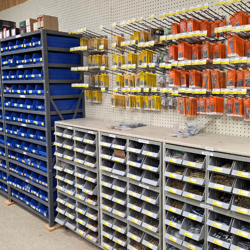 Shelving units filled with various tools and hardware in a store. Blue bins on the left, with yellow and orange packaged items hanging on hooks above.
