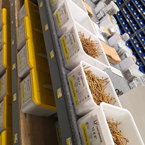 Bins with yellow lids, filled with golden screws, are neatly arranged on shelves. Labels detail specifications. A tidy, organized workshop scene.
