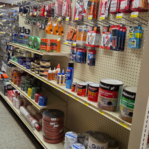 A hardware store aisle with shelves stocked with adhesives, tapes, and tools. Various containers and packets are neatly arranged, creating an organized and practical atmosphere.