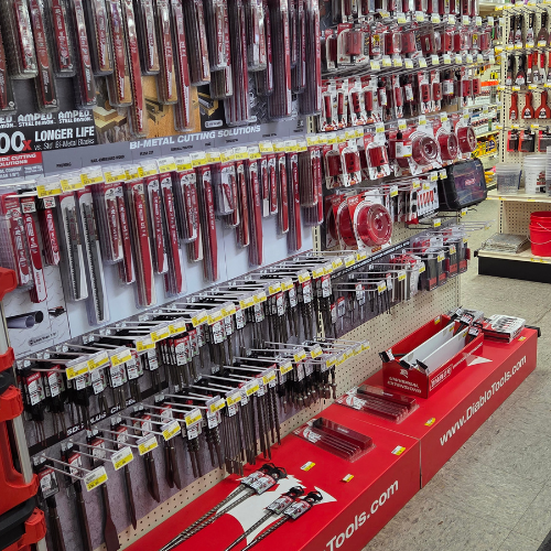 Shelves filled with an extensive variety of red and silver saw blades and cutting tools in a hardware store. The display is neat and organized.