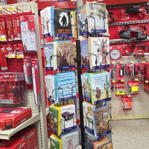 A display of colorful greeting cards stands amidst a hardware store aisle, surrounded by red tools and equipment. The scene feels vibrant and eclectic.